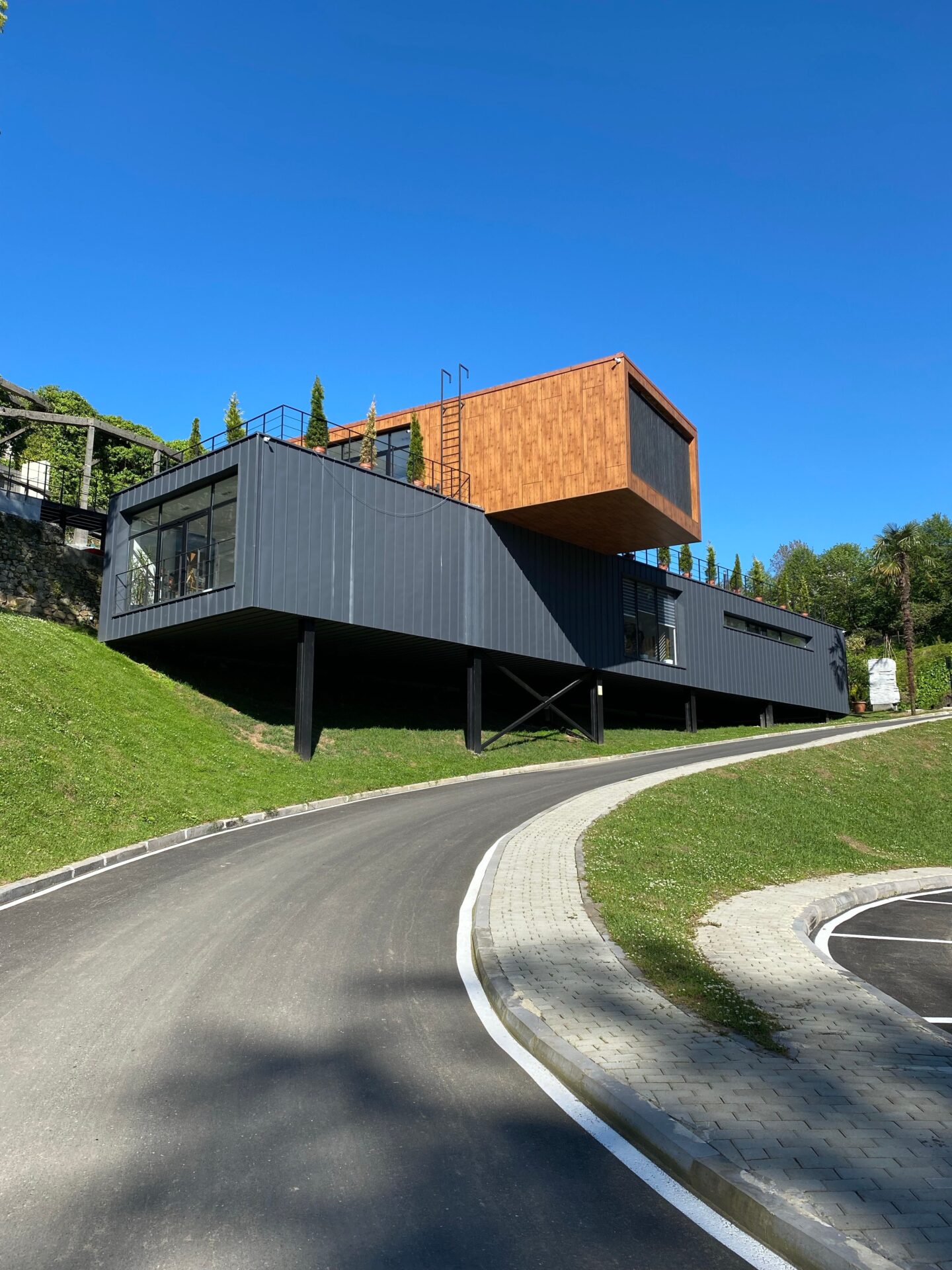 Modern steel building with black metal cladding and a cantilevered wood-accented upper level, elevated on steel supports beside a curved driveway under a clear blue sky.