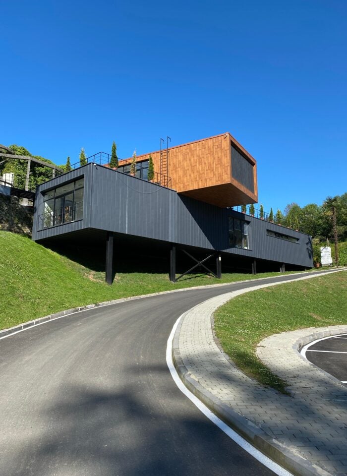 Modern steel building with black metal cladding and a cantilevered wood-accented upper level, elevated on steel supports beside a curved driveway under a clear blue sky.