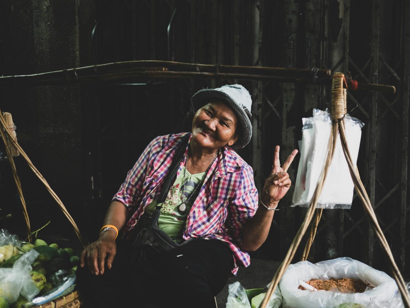 Smiling street market vendor in Thailand making a peace sign while sitting beside fresh produce.
