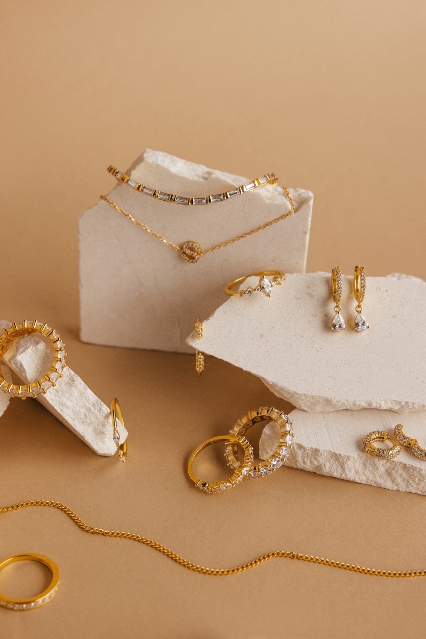 A flat lay of elegant gold jewelry displayed on white stone blocks against a warm beige background. The arrangement includes rings, hoop earrings, drop earrings, necklaces, bracelets, and a chain, many featuring clear gemstones, creating a sophisticated and minimalist luxury presentation.