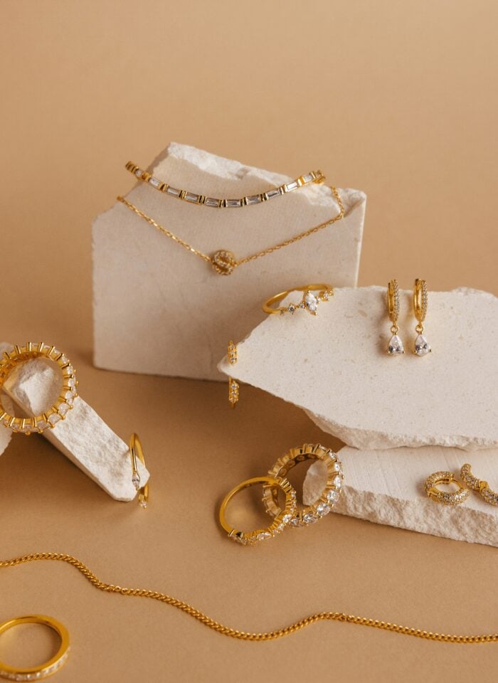 A flat lay of elegant gold jewelry displayed on white stone blocks against a warm beige background. The arrangement includes rings, hoop earrings, drop earrings, necklaces, bracelets, and a chain, many featuring clear gemstones, creating a sophisticated and minimalist luxury presentation.