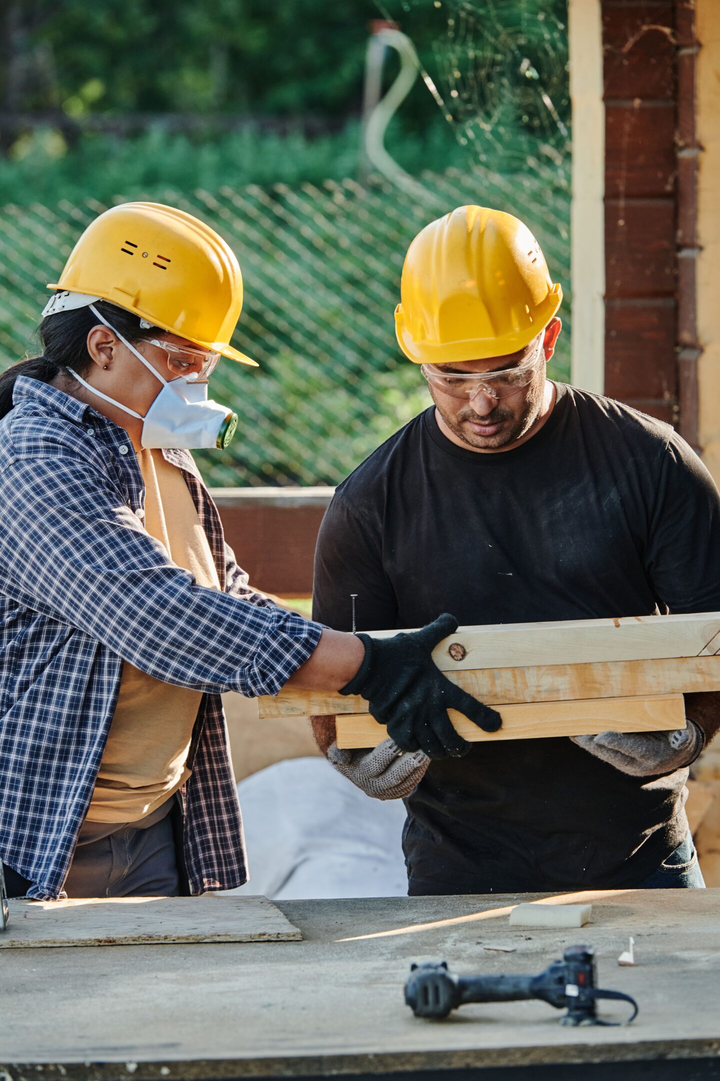 Two local construction contractors wearing hard hats and safety gear carry wooden boards during a home renovation project.