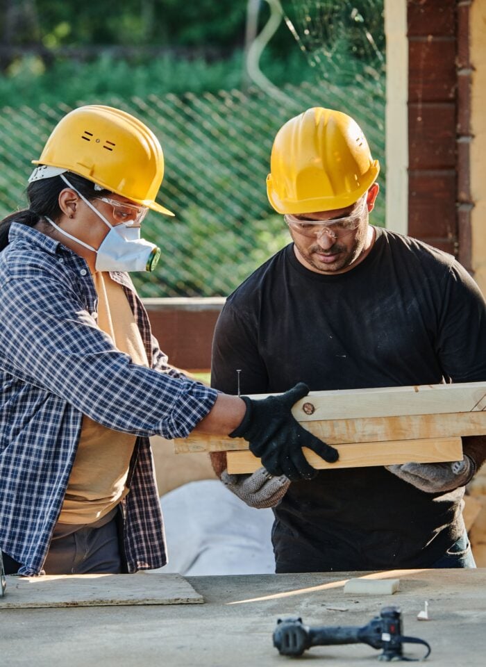 Two local construction contractors wearing hard hats and safety gear carry wooden boards during a home renovation project.