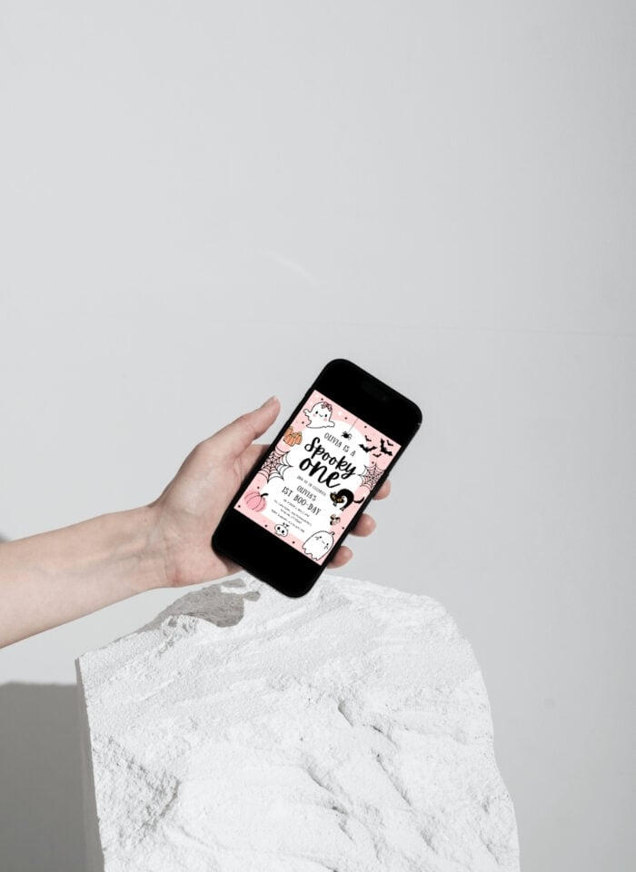 Person holding a smartphone displaying a pink digital Halloween first birthday invitation, illustrating the convenience of digital invitations for modern celebrations.