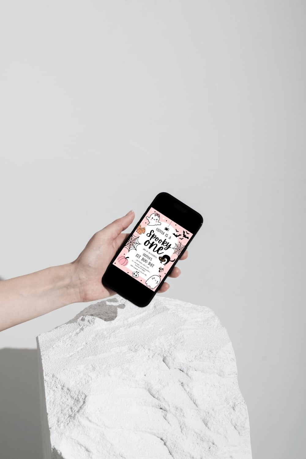 Person holding a smartphone displaying a pink digital Halloween first birthday invitation, illustrating the convenience of digital invitations for modern celebrations.