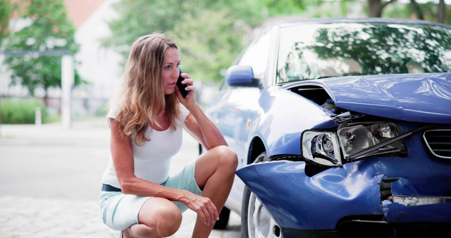 Woman using her mobile phone beside a blue car with front-end collision damage after a road traffic accident.