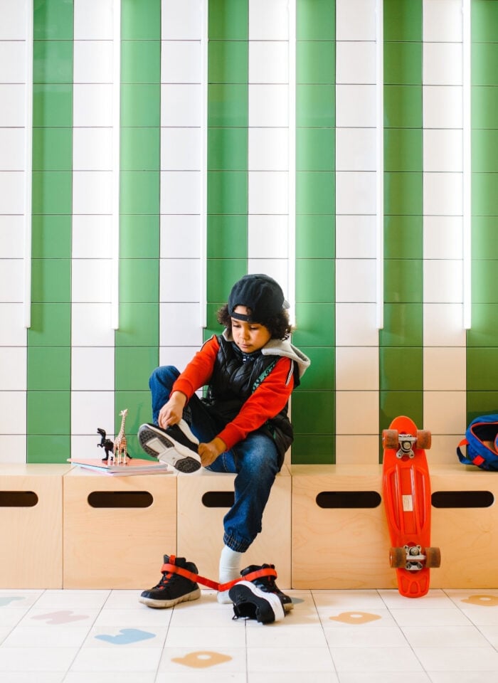 Child putting on sneakers in a school hallway with a backpack and skateboard nearby.