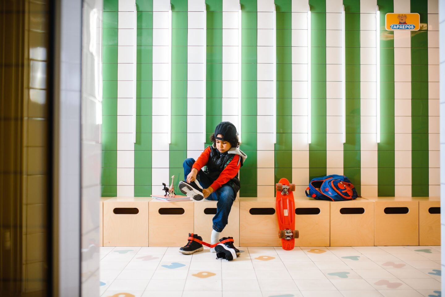 Child putting on sneakers in a school hallway with a backpack and skateboard nearby.