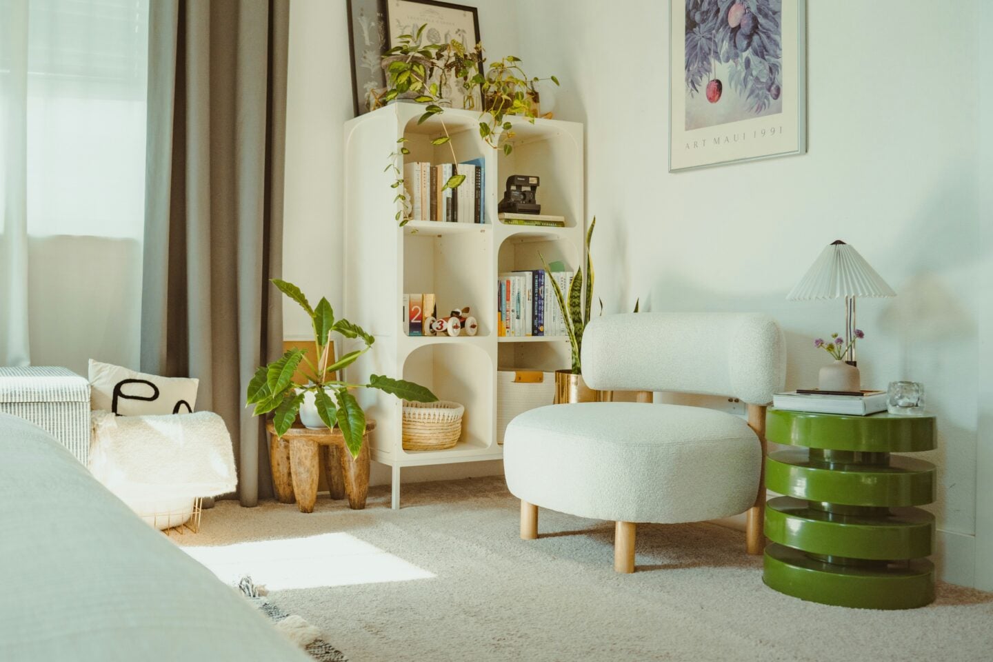 Cozy reading nook with a cream armchair, white bookshelf, indoor plants, green side table and framed botanical wall art.