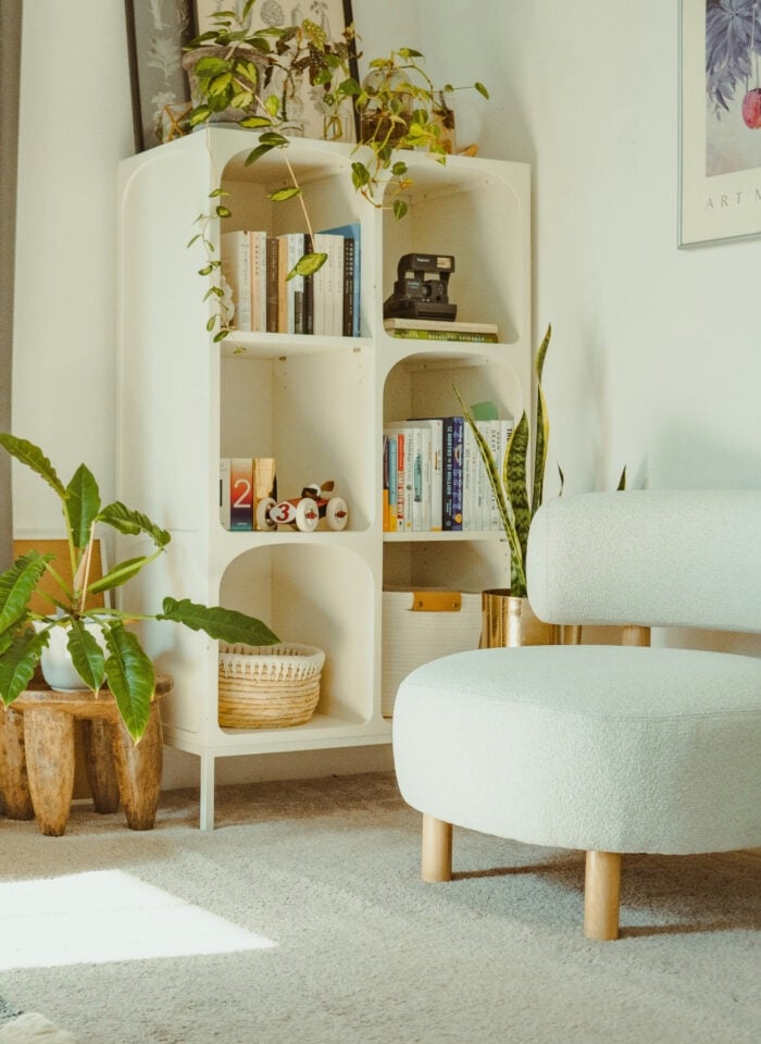 Cozy reading nook with a cream armchair, white bookshelf, indoor plants, green side table and framed botanical wall art.