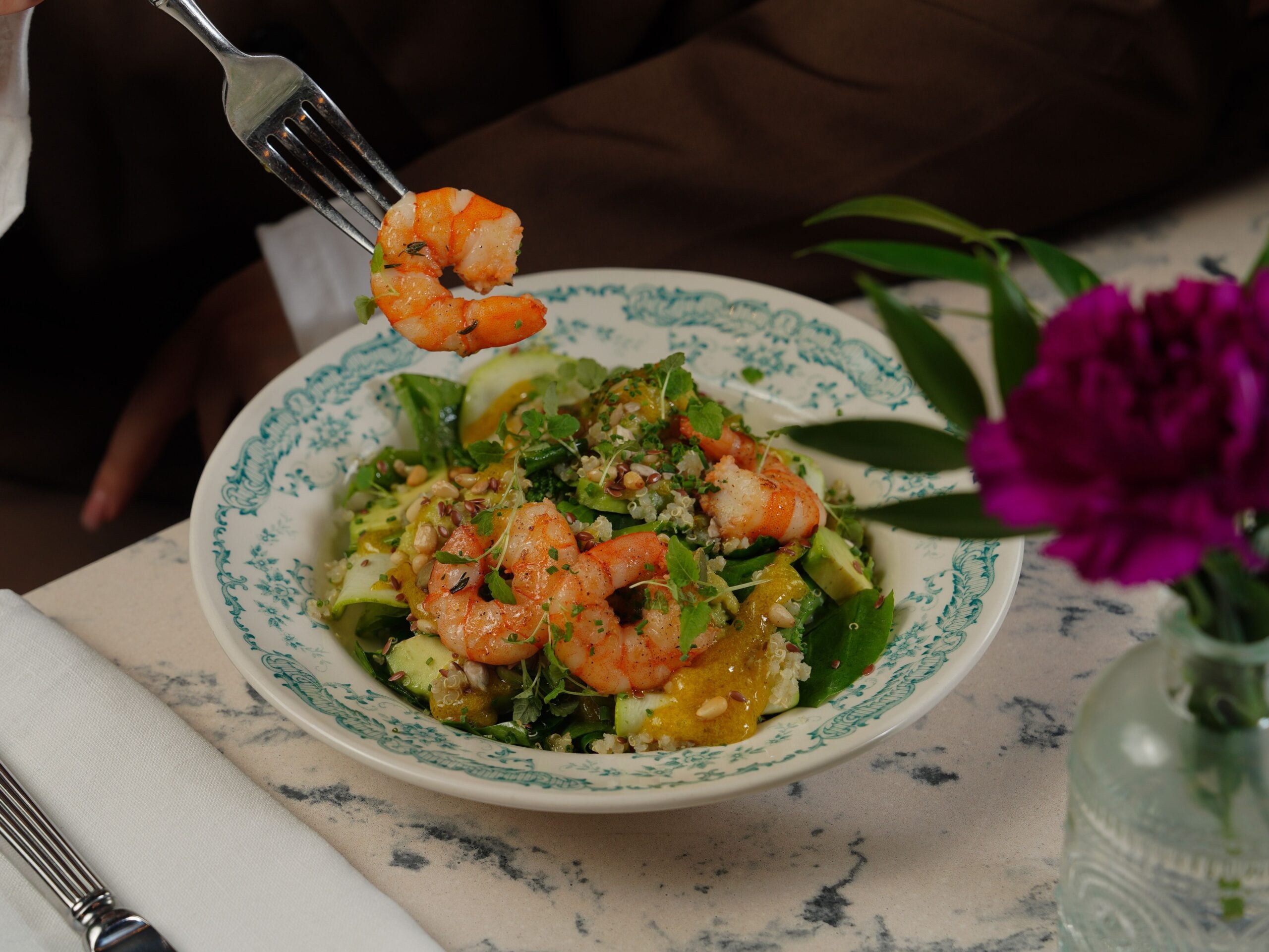 Bowl of fresh shrimp salad with leafy greens, cucumber ribbons, quinoa, citrus segments, seeds, and herbs, with a fork holding a cooked shrimp above the plate.