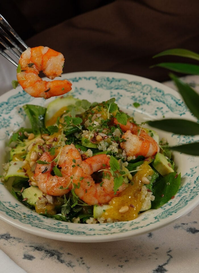 Bowl of fresh shrimp salad with leafy greens, cucumber ribbons, quinoa, citrus segments, seeds, and herbs, with a fork holding a cooked shrimp above the plate.