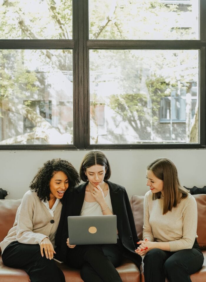 Three women sitting on a sofa looking at a laptop together.