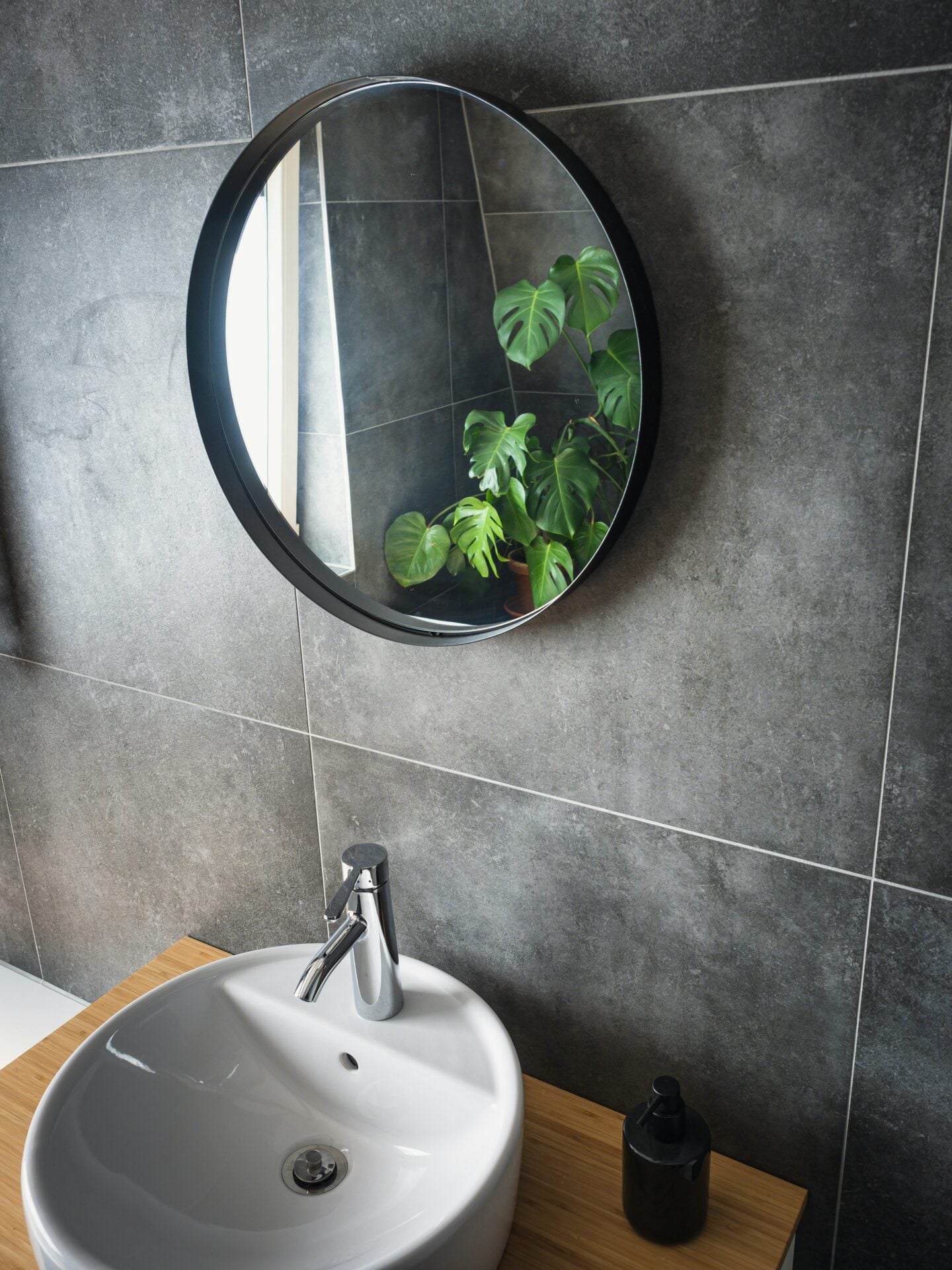 Modern bathroom with large grey wall tiles, a round black-framed mirror, white basin and wooden vanity.