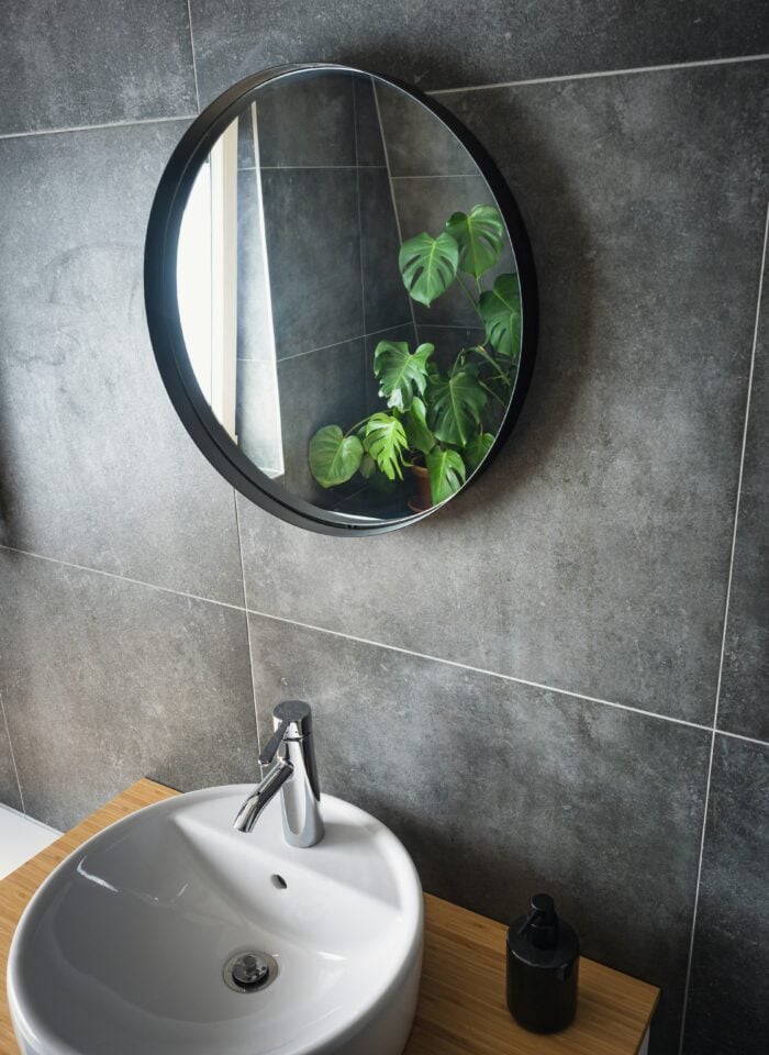 Modern bathroom with large grey wall tiles, a round black-framed mirror, white basin and wooden vanity.