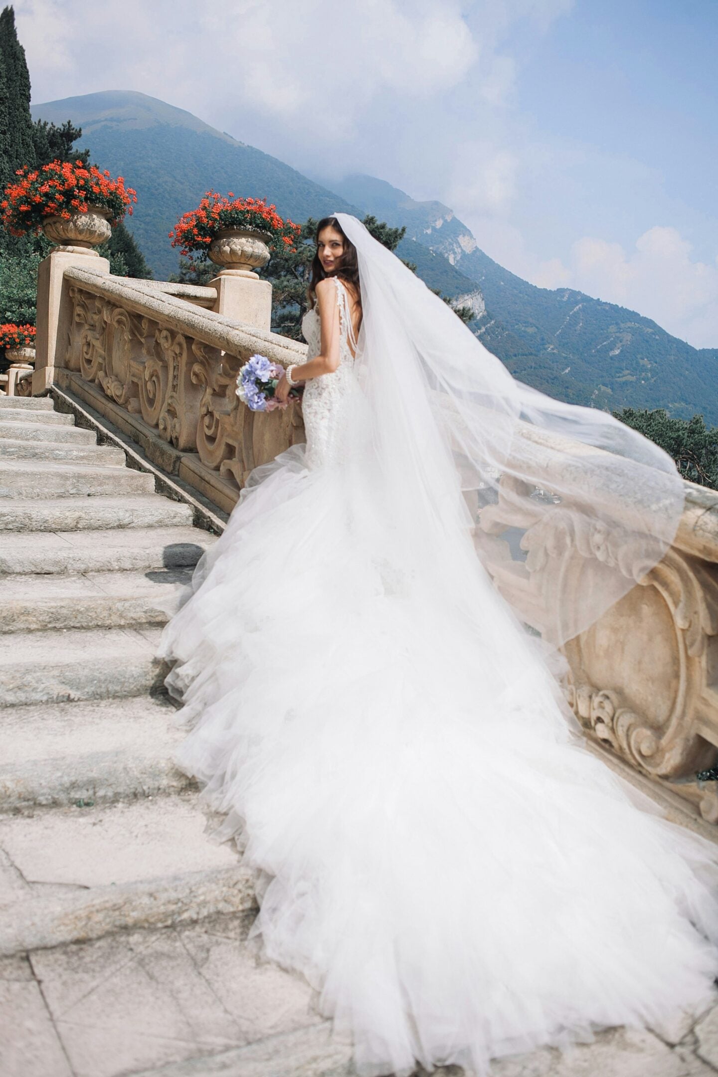 Bride wearing a white wedding dress and long veil on stone steps with mountains in the background.