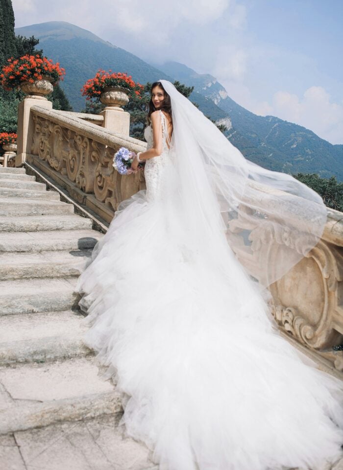 Bride wearing a white wedding dress and long veil on stone steps with mountains in the background.