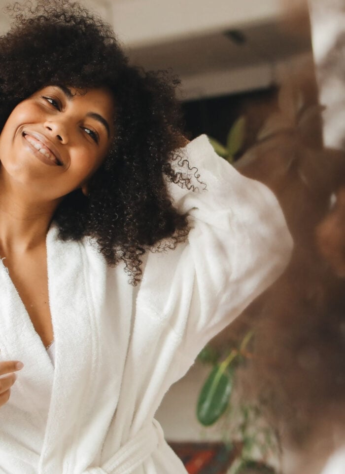 Smiling woman in a white bathrobe looking at herself in the mirror as part of a self-care routine.
