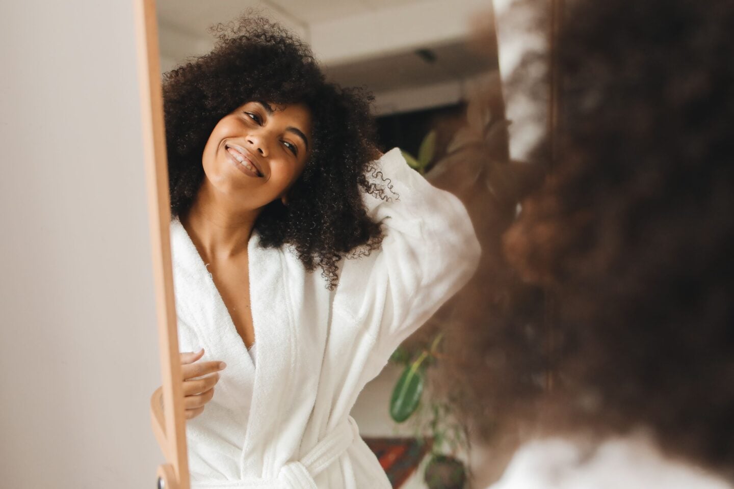 Smiling woman in a white bathrobe looking at herself in the mirror as part of a self-care routine.