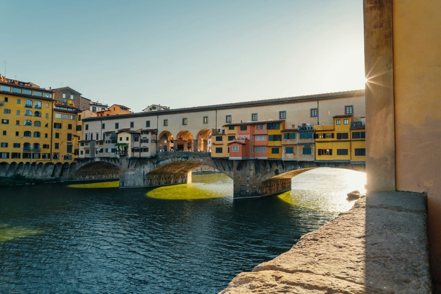 Ponte Vecchio bridge spanning the Arno River in Florence, Italy, with its historic buildings illuminated by the afternoon sun.