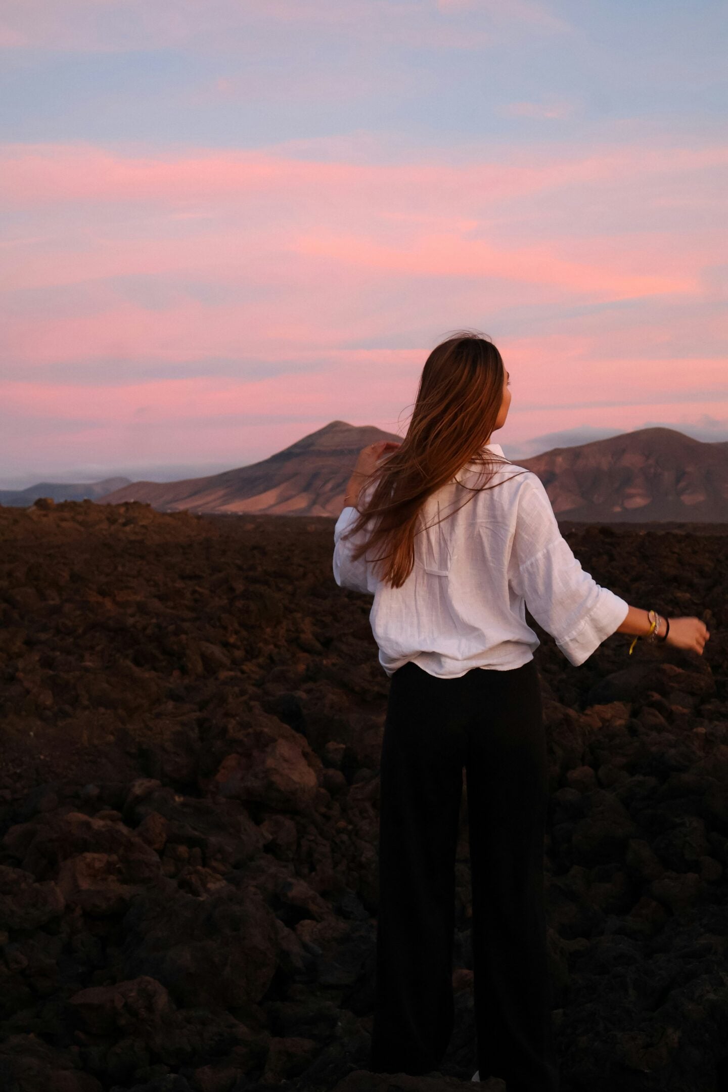 Woman standing on a rocky volcanic landscape with mountains under a pink sunset sky.