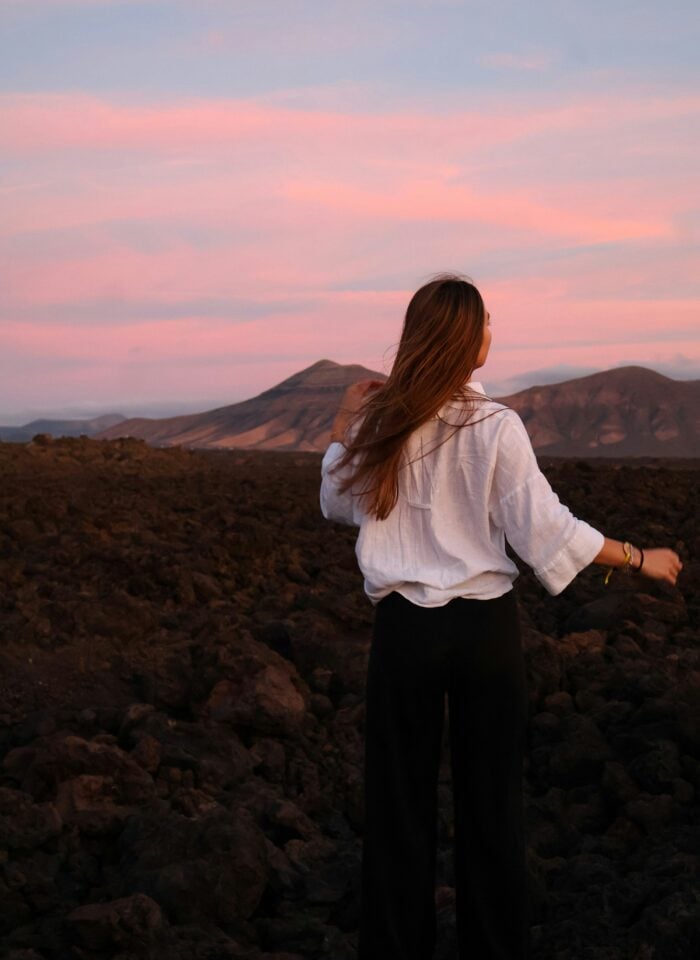 Woman standing on a rocky volcanic landscape with mountains under a pink sunset sky.