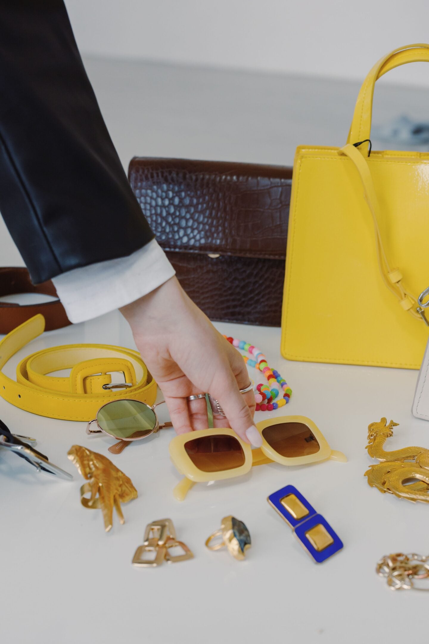 Woman selecting sunglasses from a display of colorful fashion accessories, including a yellow handbag, belt, jewelry, and eyewear.