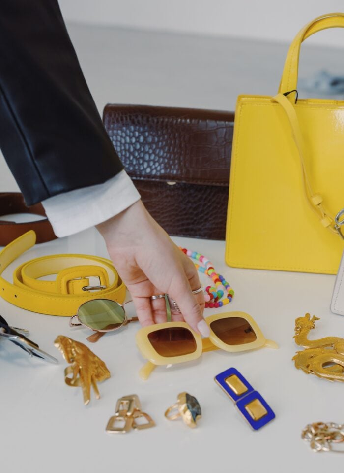 Woman selecting sunglasses from a display of colorful fashion accessories, including a yellow handbag, belt, jewelry, and eyewear.