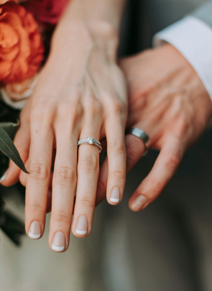 Bride and groom showing their wedding and engagement rings beside a bouquet of orange flowers and greenery.