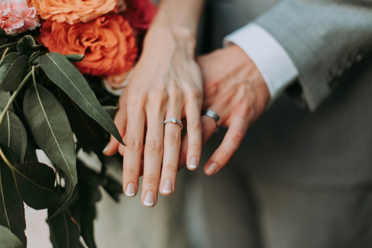 Bride and groom showing their wedding and engagement rings beside a bouquet of orange flowers and greenery.