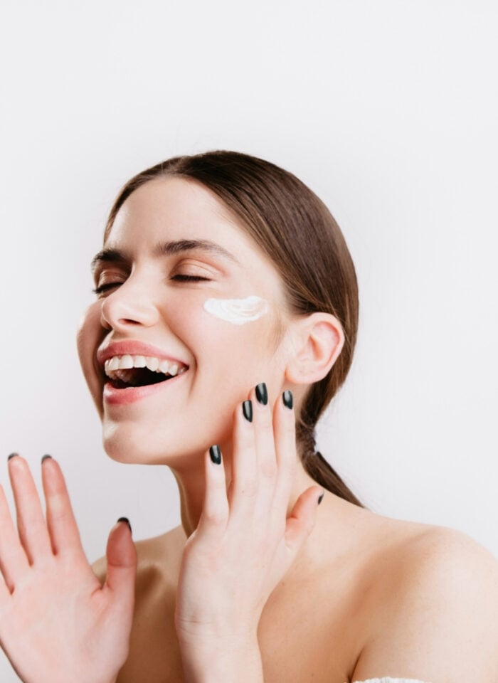 Smiling woman with her eyes closed applying face cream to her cheek as part of a modern skincare and self-care routine against a soft neutral background.