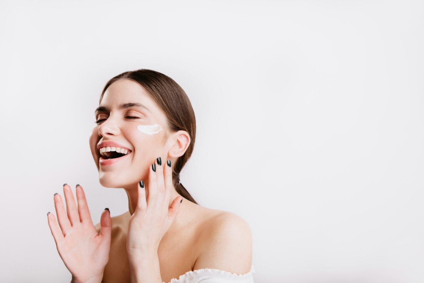 Smiling woman with her eyes closed applying face cream to her cheek as part of a modern skincare and self-care routine against a soft neutral background.