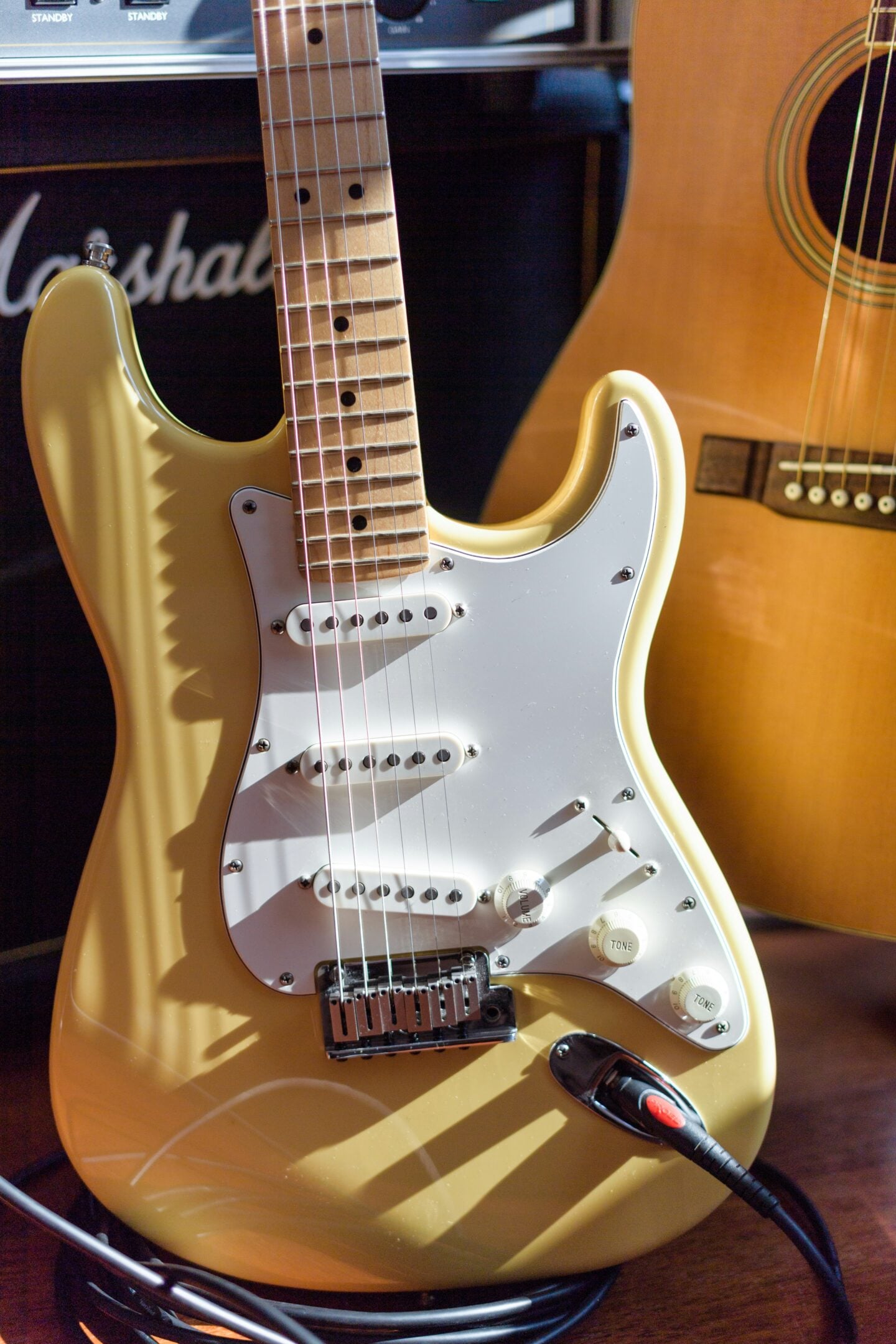 Close-up of a gold electric guitar with a white pickguard, with an acoustic guitar and amplifier in the background.