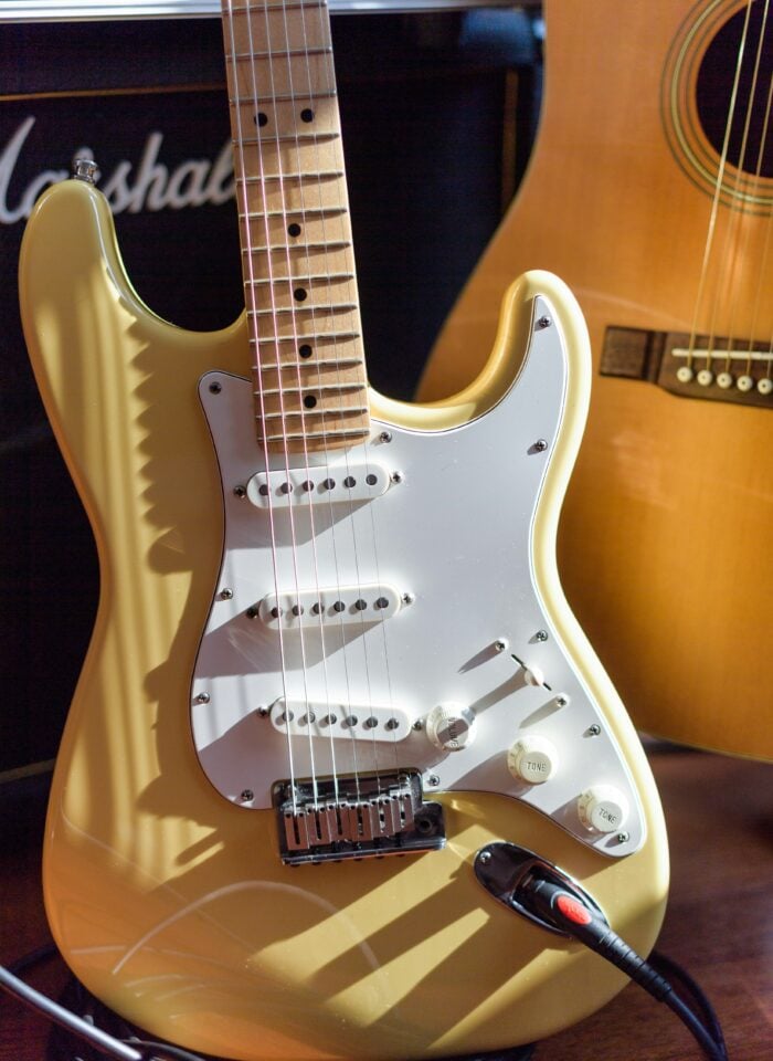 Close-up of a gold electric guitar with a white pickguard, with an acoustic guitar and amplifier in the background.