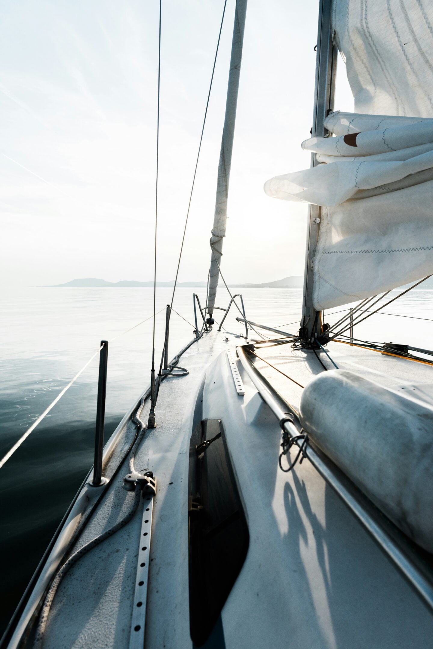 View from the deck of a sailboat gliding across calm water at sunrise, with folded sails, rigging, and distant mountains visible on the horizon.