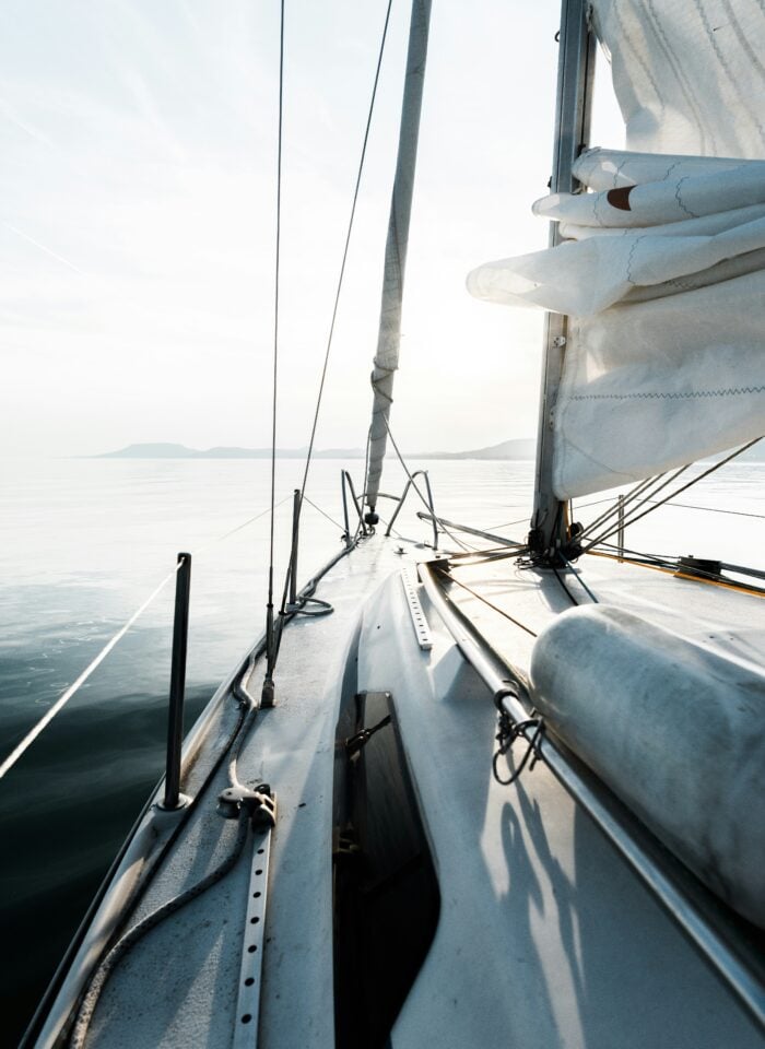 View from the deck of a sailboat gliding across calm water at sunrise, with folded sails, rigging, and distant mountains visible on the horizon.