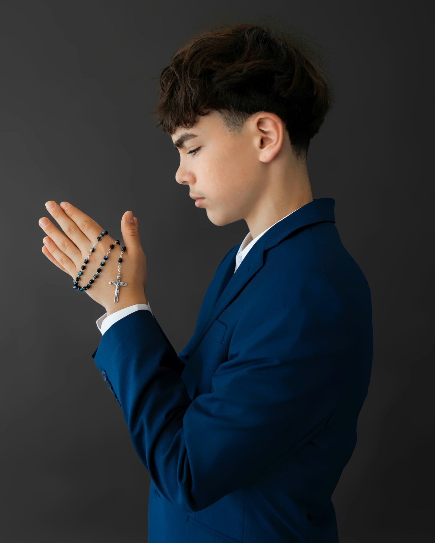 Young boy in a blue suit holding rosary beads with a cross while praying.