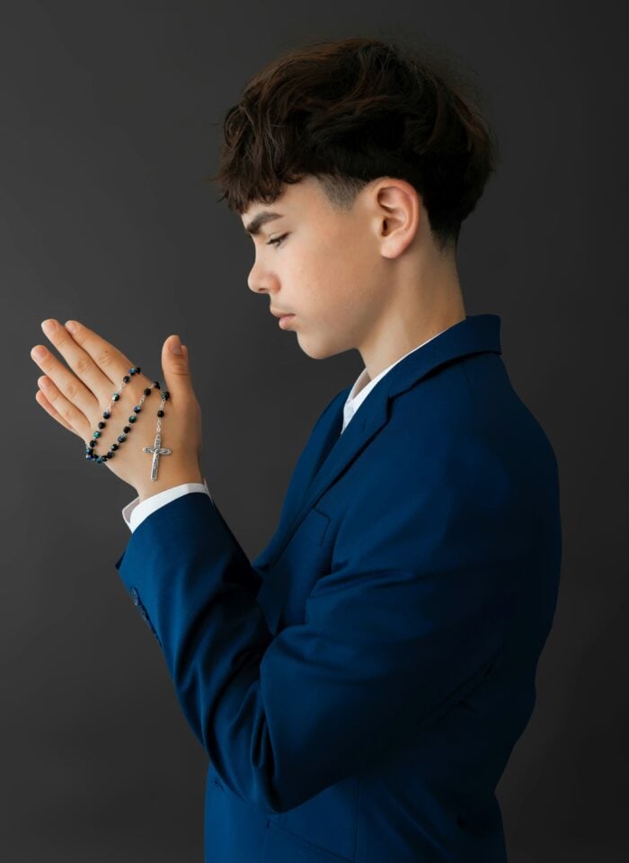 Young boy in a blue suit holding rosary beads with a cross while praying.