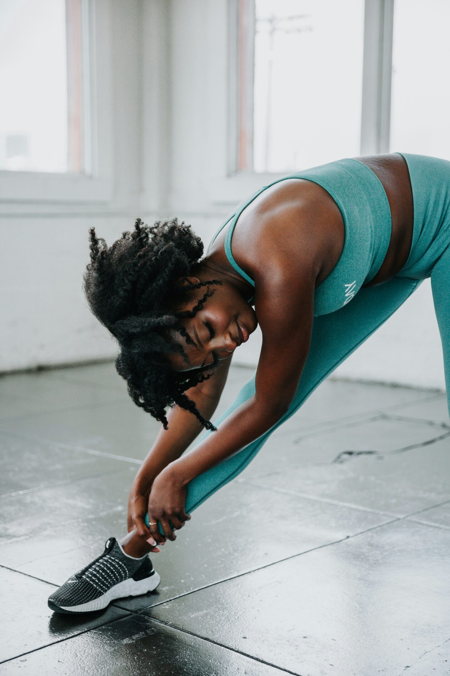 A woman wearing a teal sports bra and matching high-waisted leggings stretches in a bright fitness studio. She is leaning deeply to one side with her upper body bent over her extended leg, reaching down with both hands to hold her ankle. She wears black-and-white athletic shoes and has curly dark hair falling forward as she stretches. Large windows fill the background with natural light, while the polished grey tiled floor reflects some of the light.