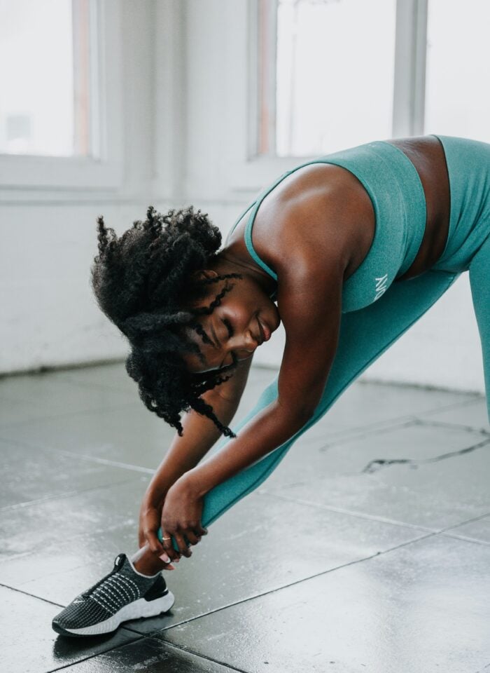 A woman wearing a teal sports bra and matching high-waisted leggings stretches in a bright fitness studio. She is leaning deeply to one side with her upper body bent over her extended leg, reaching down with both hands to hold her ankle. She wears black-and-white athletic shoes and has curly dark hair falling forward as she stretches. Large windows fill the background with natural light, while the polished grey tiled floor reflects some of the light.