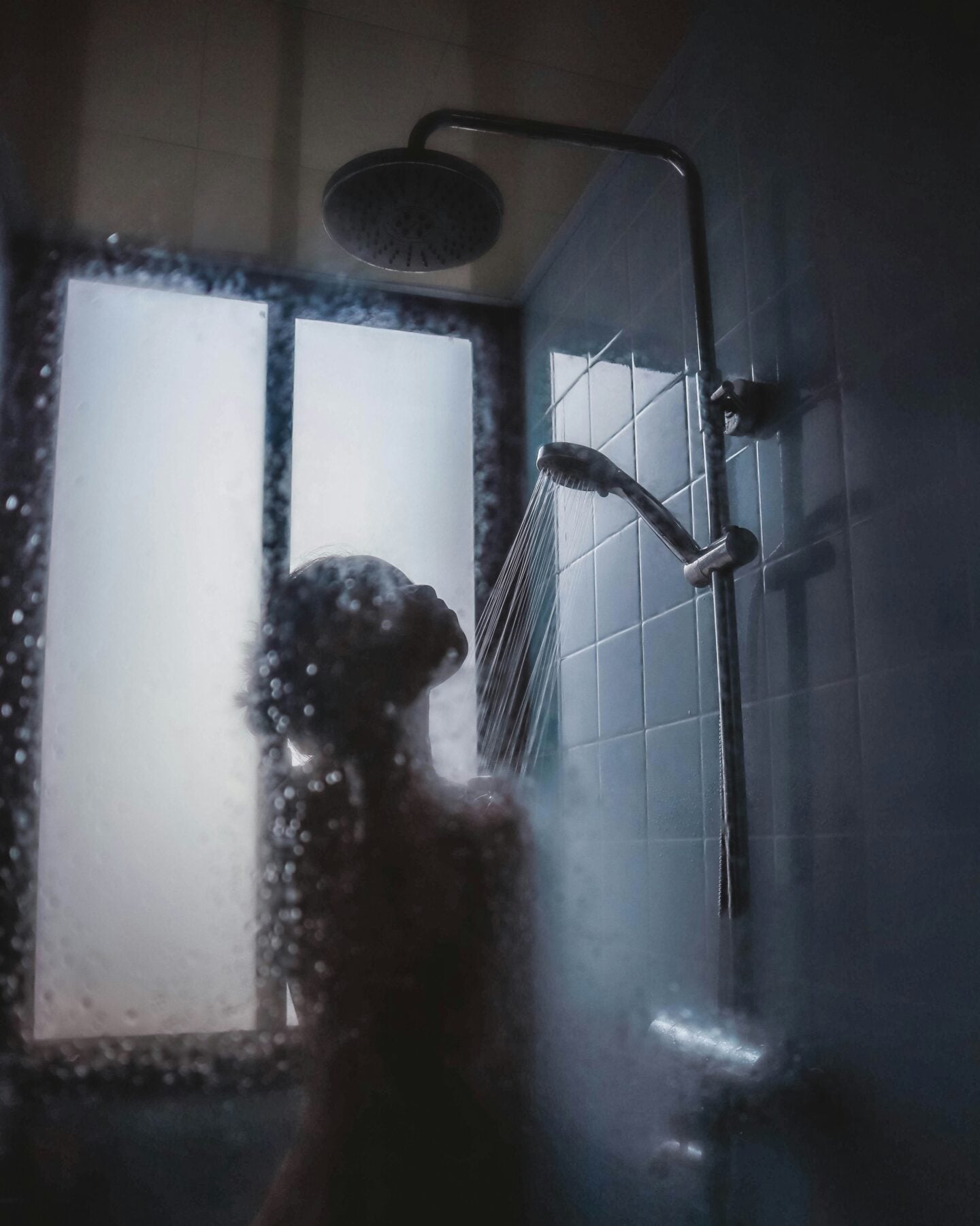 Woman washing her hair under a handheld shower head in a steamy bathroom.