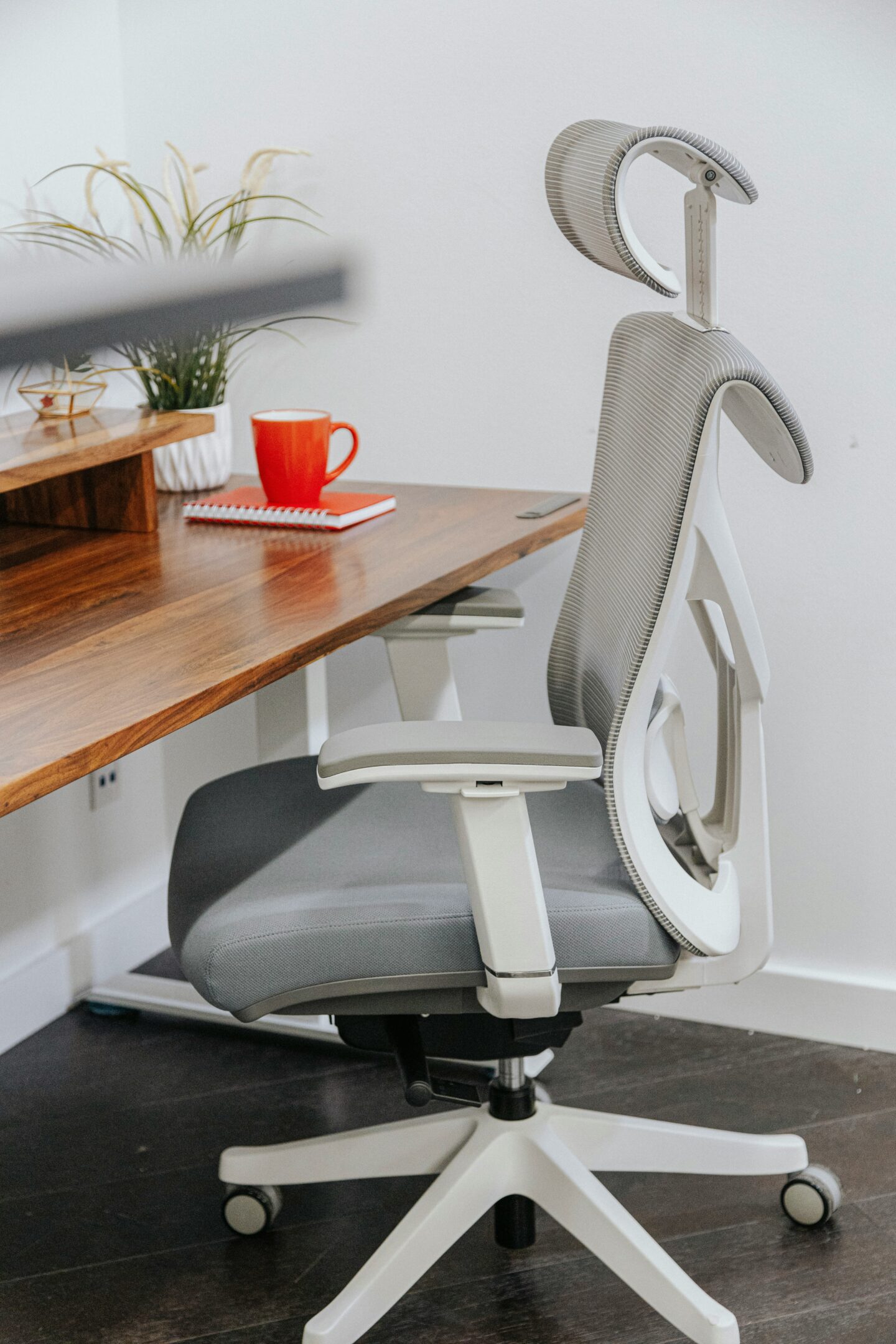 Grey ergonomic office chair with adjustable headrest, armrests and lumbar support positioned beside a wooden home office desk.