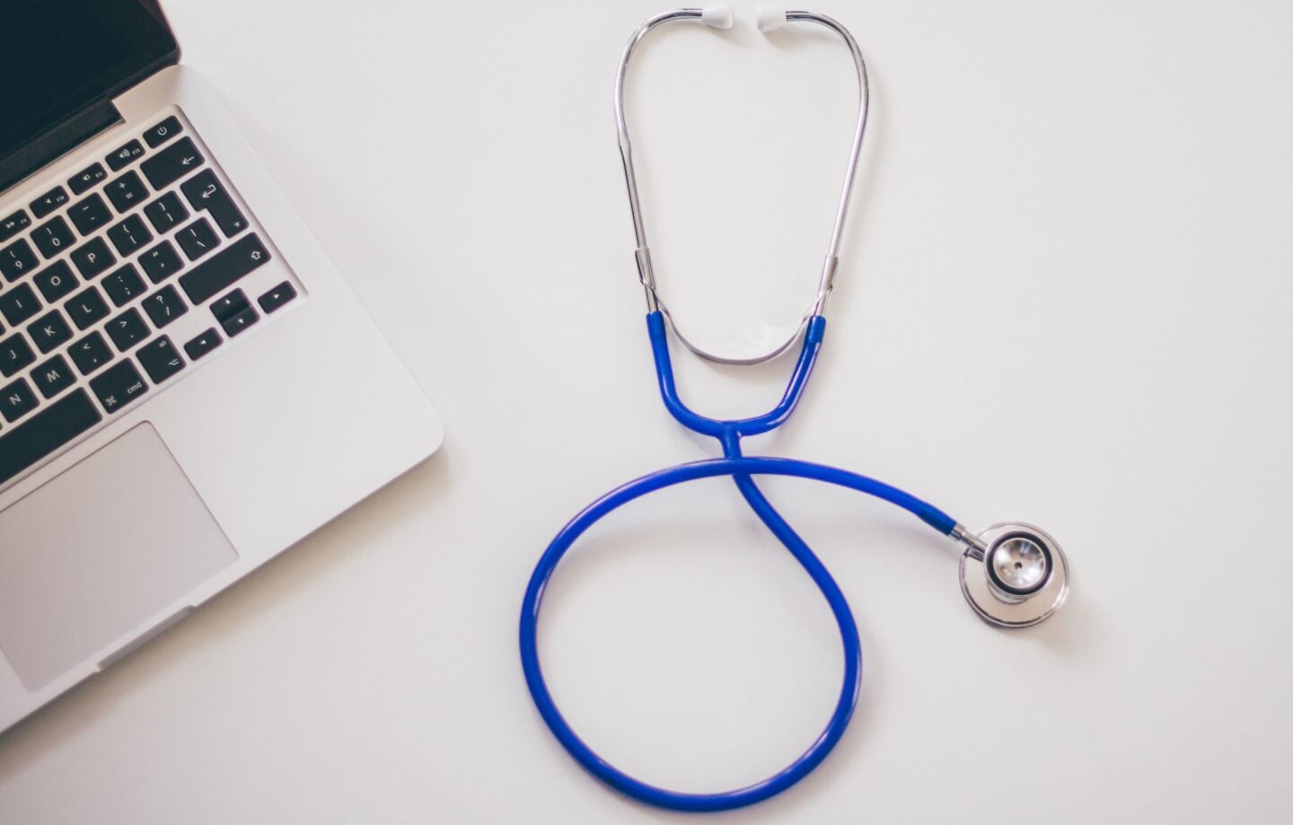 Blue stethoscope beside a laptop on a white desk, representing healthcare and clinical business management.