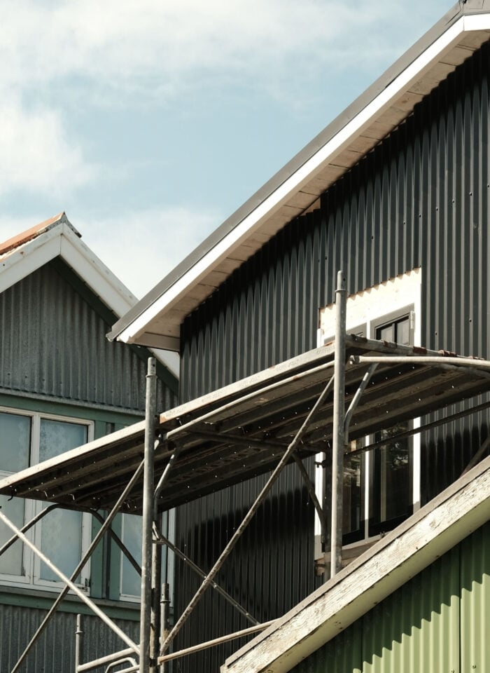 Exterior of corrugated metal houses with black and green siding, white-trimmed windows, pitched roofs and scaffolding.