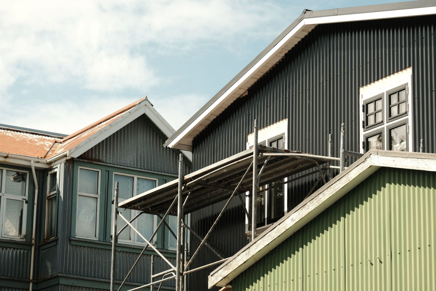 Exterior of corrugated metal houses with black and green siding, white-trimmed windows, pitched roofs and scaffolding.