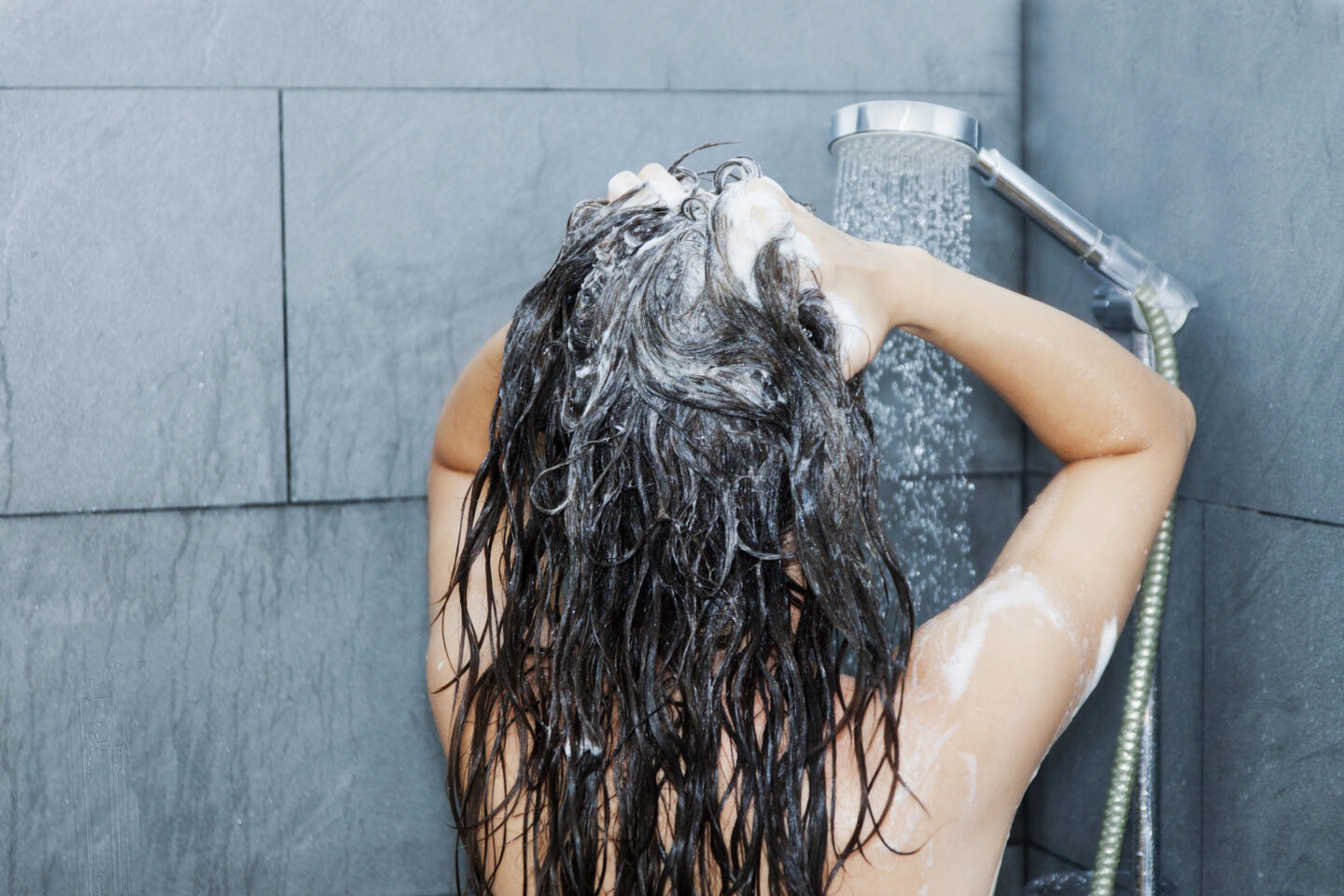 Woman washing her long dark hair with shampoo under a shower.