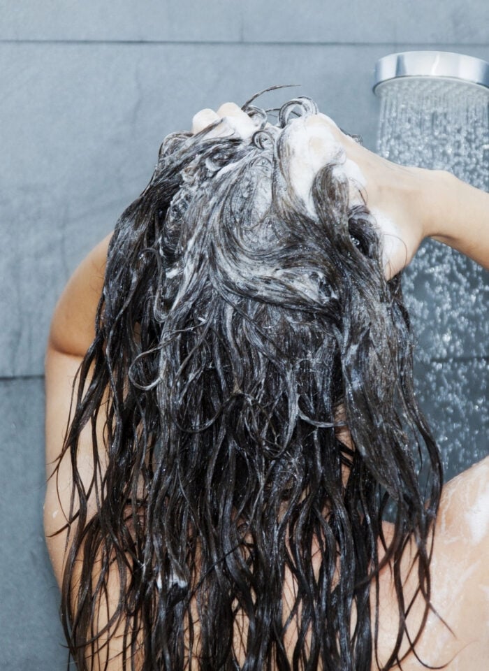 Woman washing her long dark hair with shampoo under a shower.