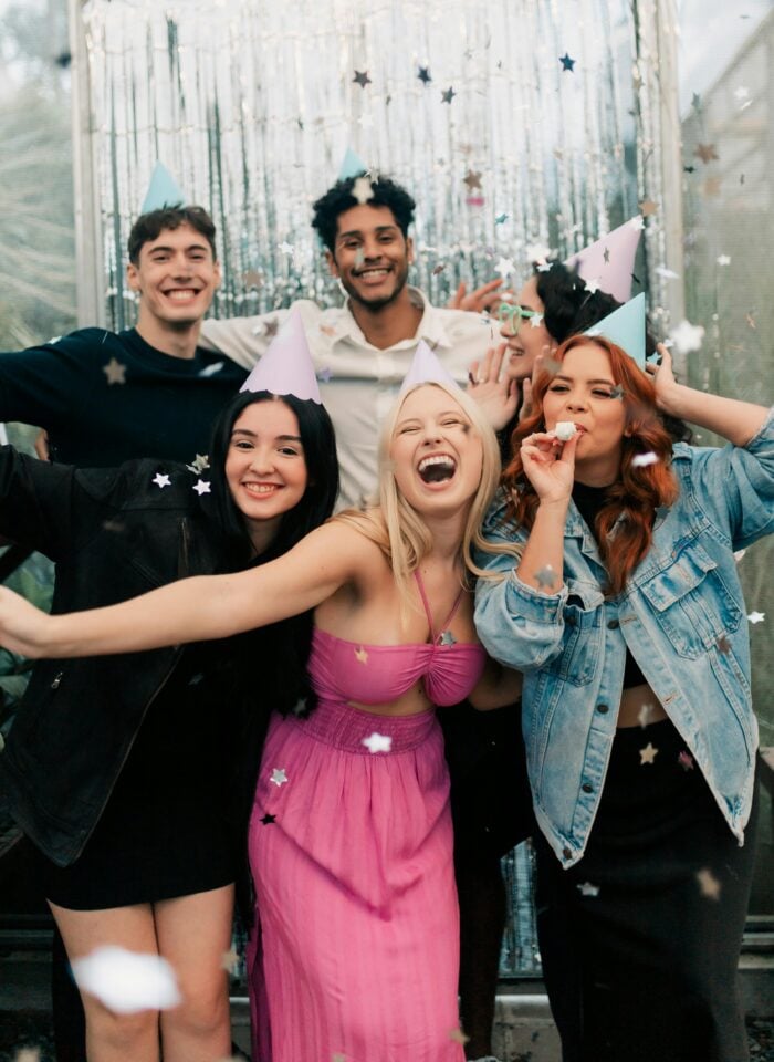 Group of friends wearing party hats smiling and posing together with confetti falling.