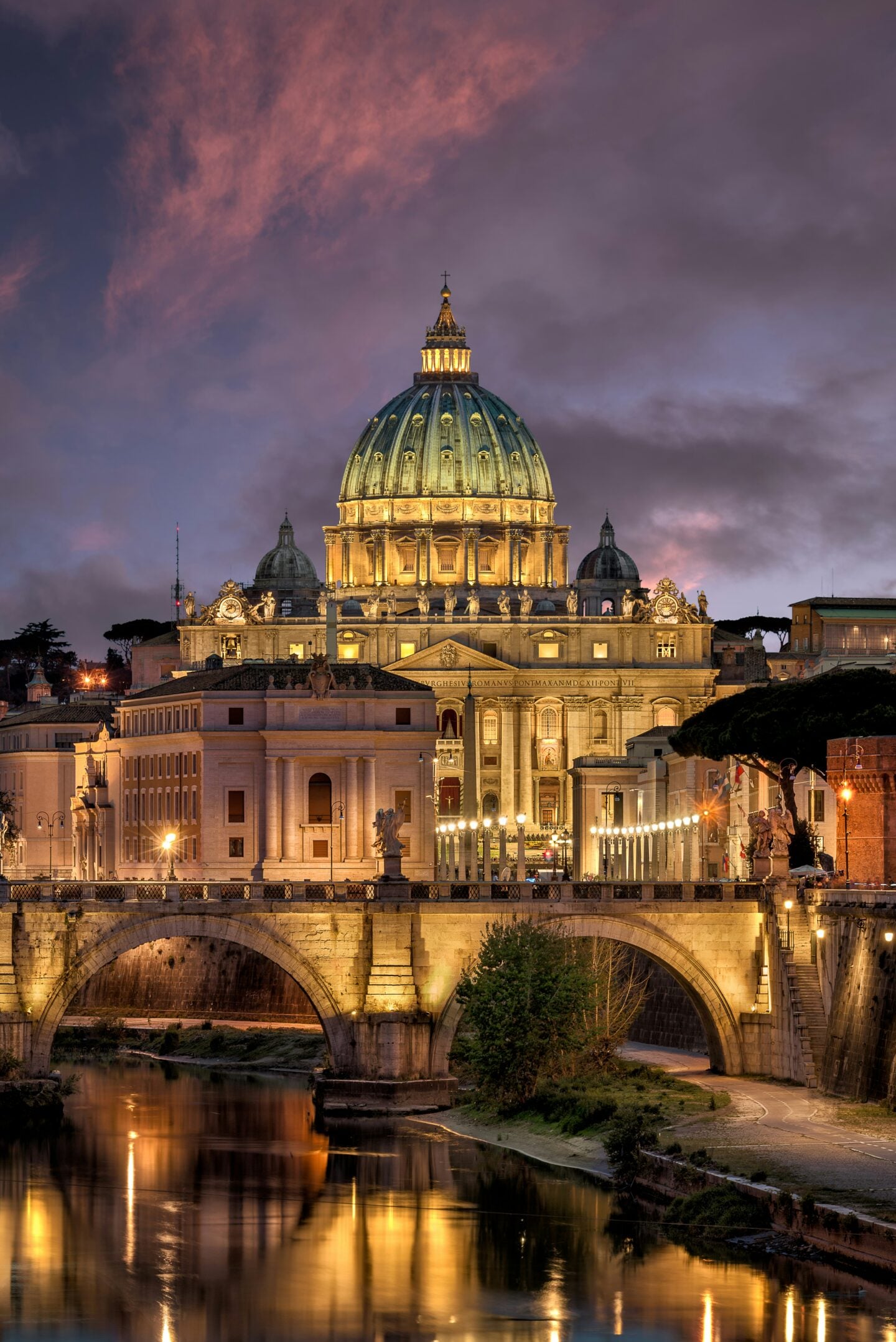 Illuminated St. Peter’s Basilica in Vatican City at dusk, viewed across the Tiber River in Rome.