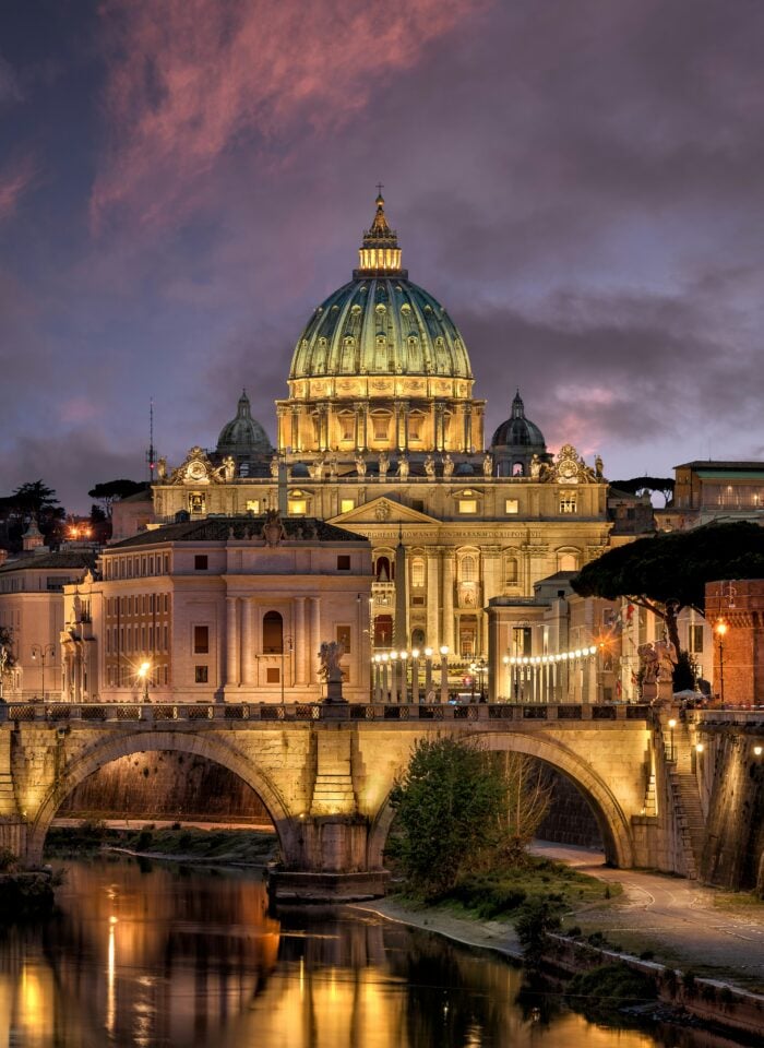Illuminated St. Peter’s Basilica in Vatican City at dusk, viewed across the Tiber River in Rome.
